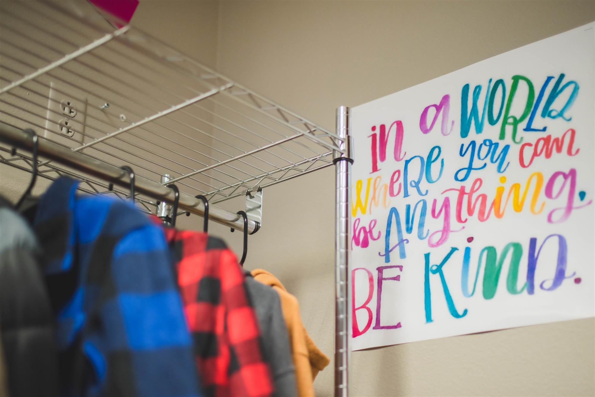 coats hang near a poster that reads "in a world where you can be anything, be kind." 