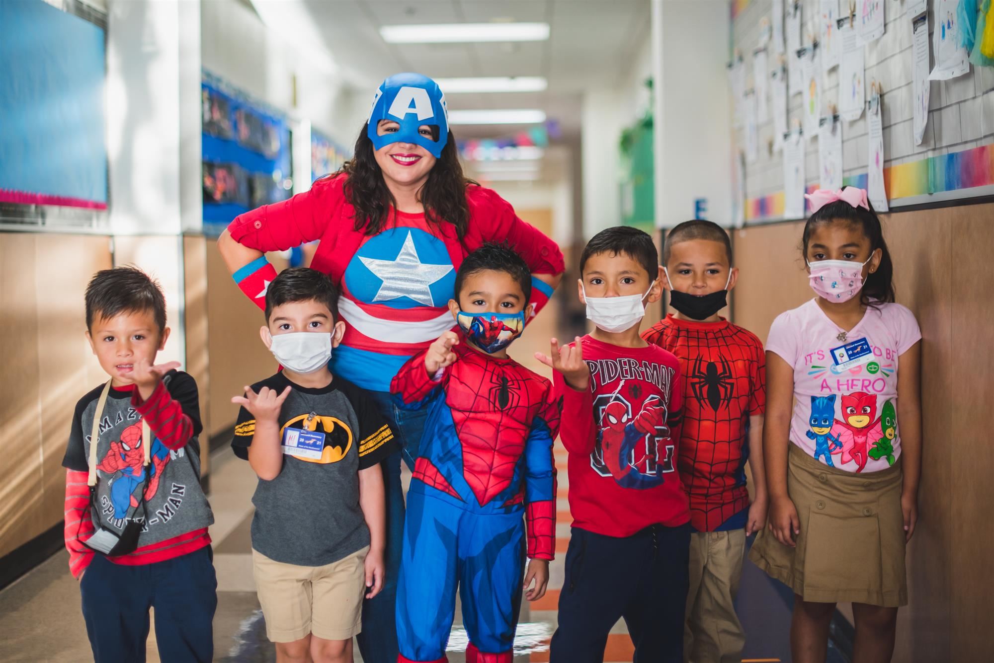 Students wear superhero costumes with teacher in hallway